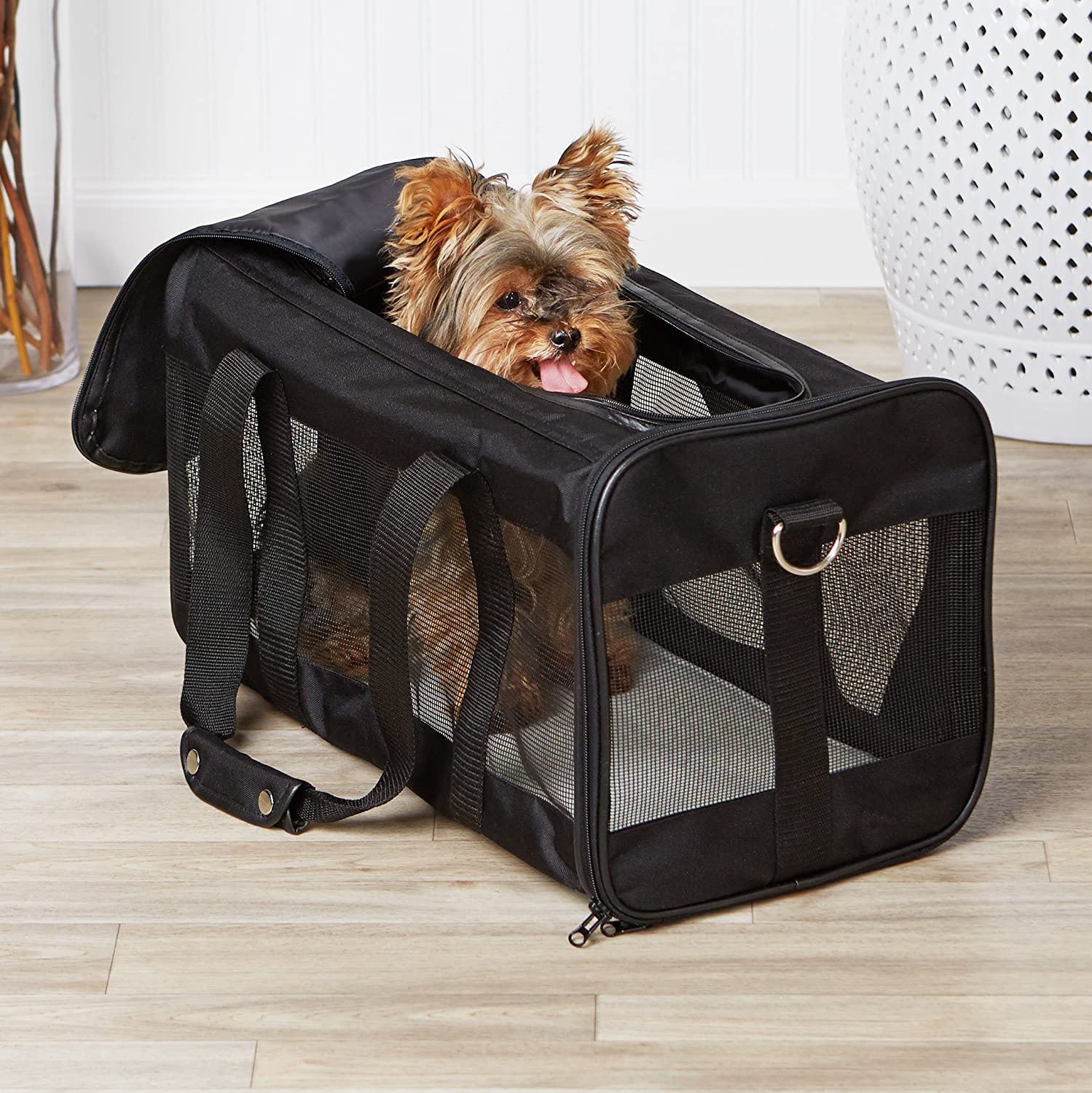 Small dog peeking out of a black pet carrier on a wooden floor.